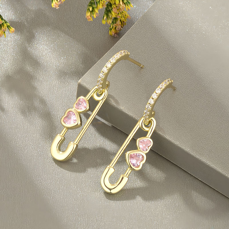 Origsoul gold heart shaped safety pin earrings with pink heart gemstones on textured background