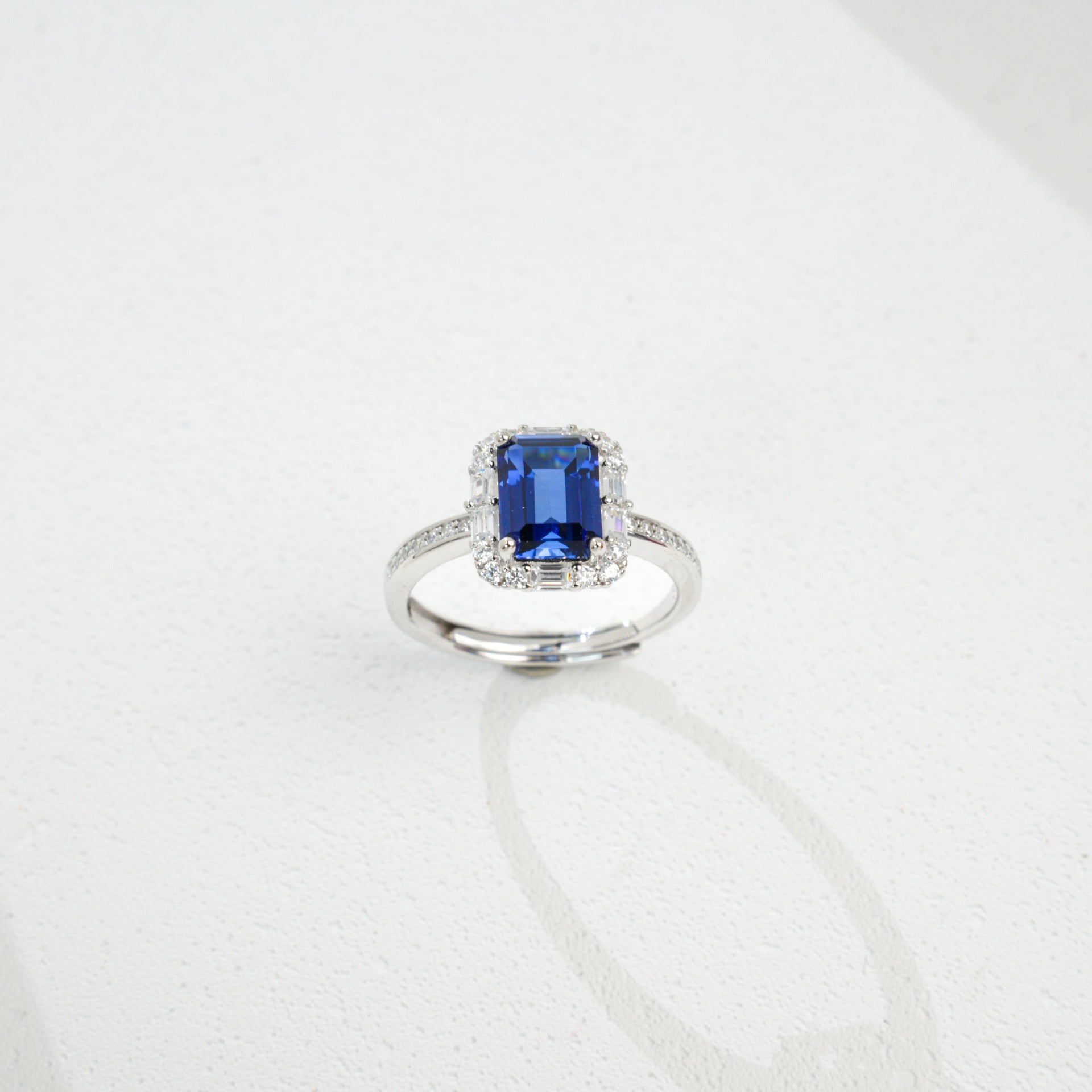 Origsoul emerald cut-engagement ring with a deep blue gemstone surrounded by small diamonds on silver band